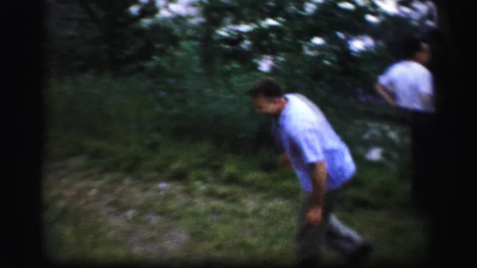 A man in a light blue shirt moves near roadside brush while another adult is partly visible at the edge of the wooded scene.
