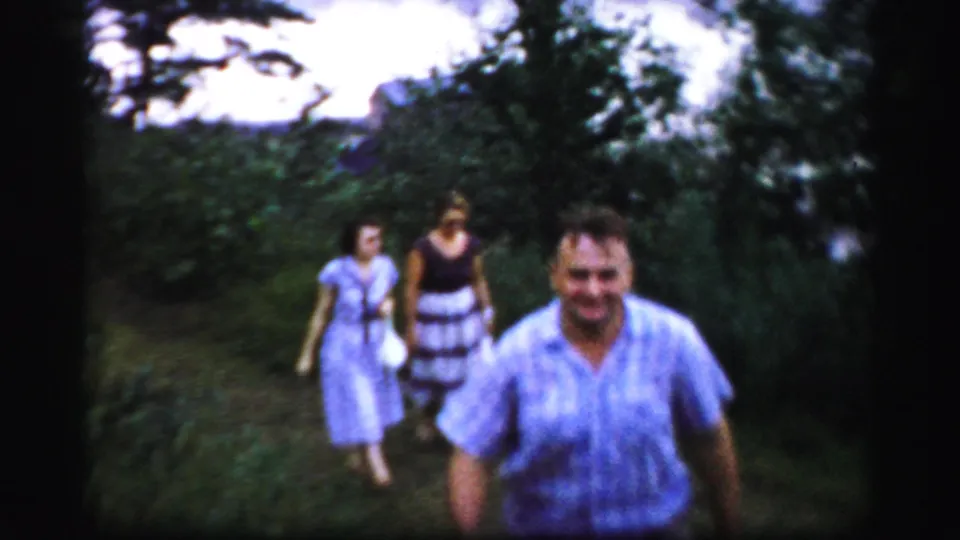 A smiling man in a light blue shirt walks close to the camera as two women in dresses follow on a shaded Albany path.