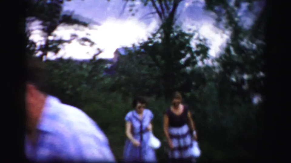The lead man is partly cropped in the foreground while two women continue walking through the leafy 1950s home-movie scene.