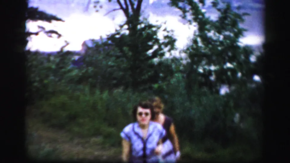 Two adult women, one in sunglasses, move closer to the camera along a green path in 1950s Albany-area home-movie footage.
