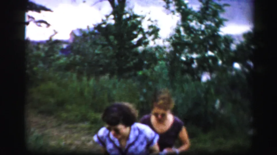 Two adult women pass near the camera while walking through dense greenery in soft 8mm footage from the Albany-area reel.