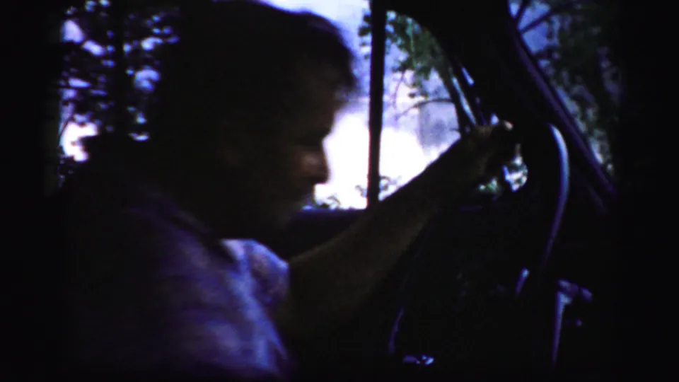 An adult man is seen in profile inside a car, framed by the windshield and steering wheel in 1950s Albany-area home-movie footage.
