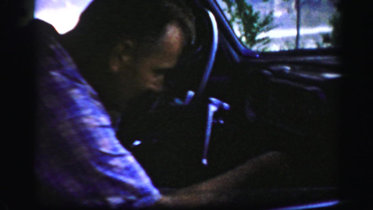 In 1956 Albany home-movie footage, a man sits in the driver's seat of a dark vintage car with window light behind him.