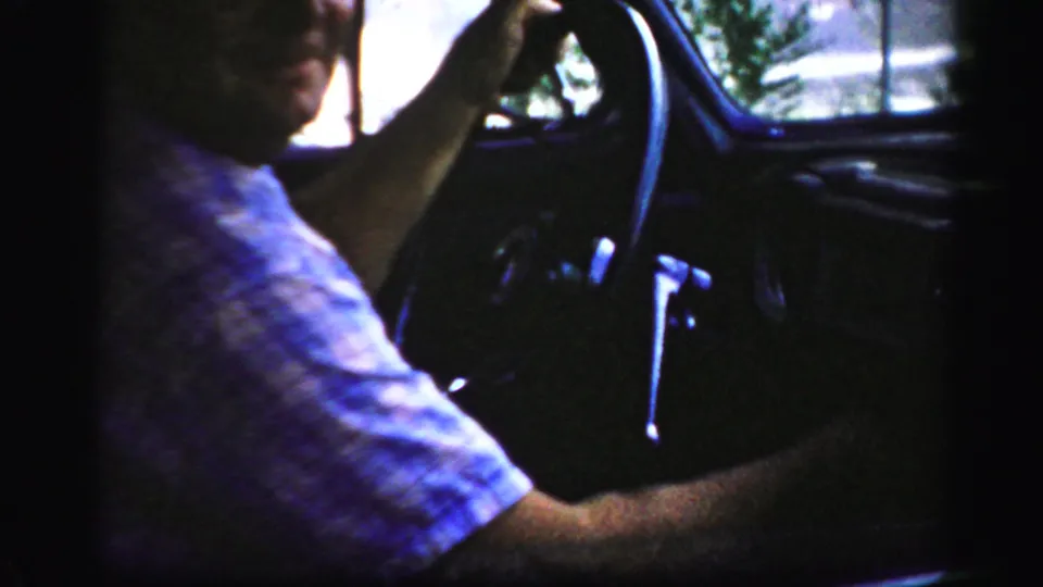 A man in a short-sleeve shirt turns partly toward the camera from the driver's seat of a 1956 Albany home movie car scene.