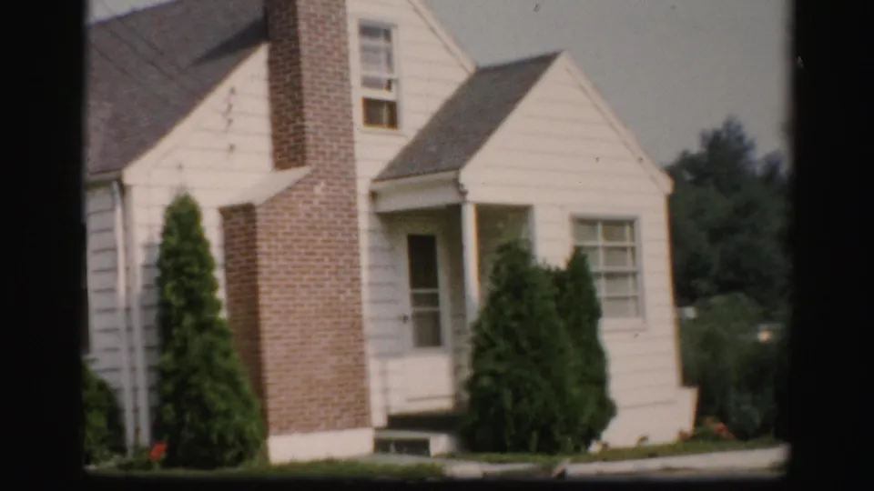 A white suburban house exterior with a brick chimney and trimmed shrubs appears in Albany-area home-movie footage from the 1950s.
