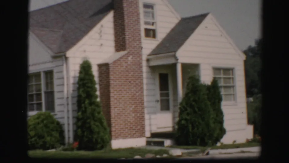 The camera holds on a white house with a brick chimney, front entry, windows, shrubs, and lawn in 1950s Albany-area footage.