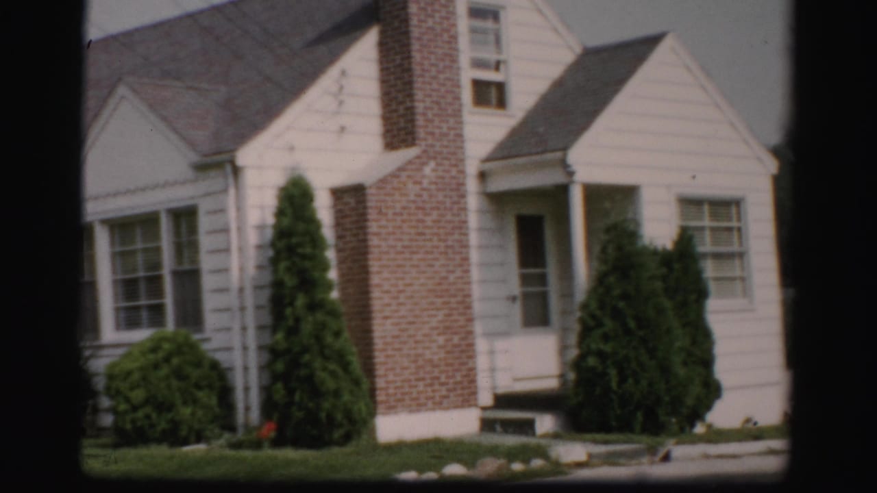 White clapboard house with a brick chimney, front windows, shrubs, lawn, and curb.