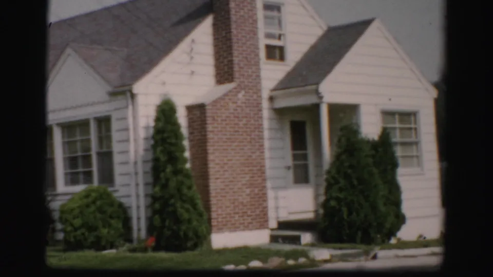 1956 Albany home-movie frame showing a white clapboard house with a brick chimney, front windows, shrubs, and green lawn.