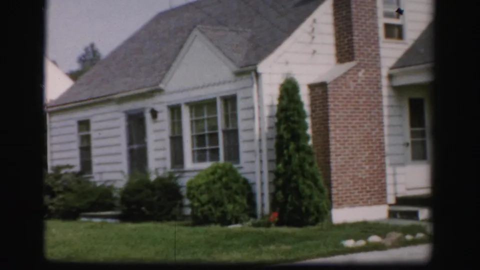 1956 Albany home-movie frame showing an angled view of a white house with gray roof, brick chimney, front windows, and lawn.
