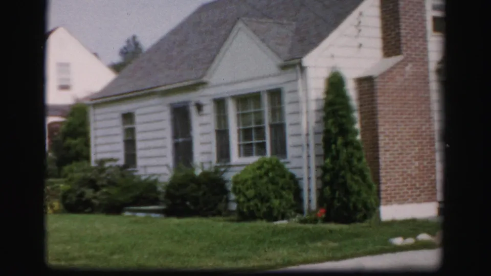 1956 Albany frame showing the white house, brick chimney, trimmed front landscaping, green lawn, and part of a neighboring house.