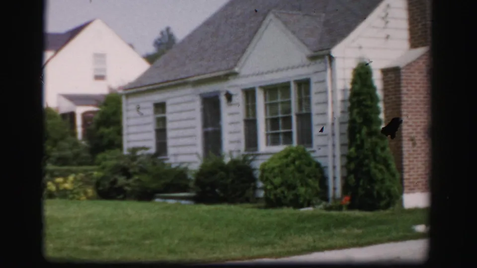 1950s Albany home-movie frame showing a white suburban house, green lawn, shrubs, and nearby residences.