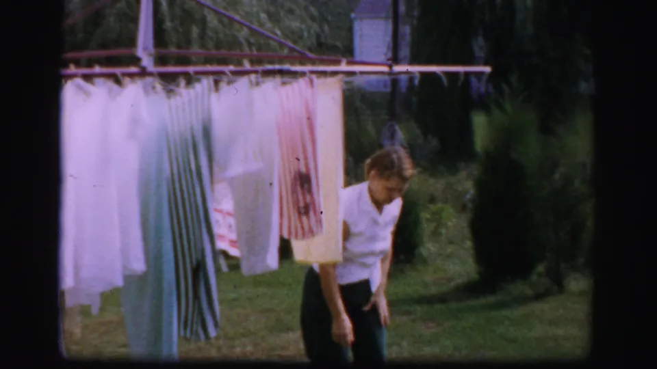 A woman in a white shirt stands near sheets and striped laundry on an outdoor clothesline in a 1950s Albany backyard scene.