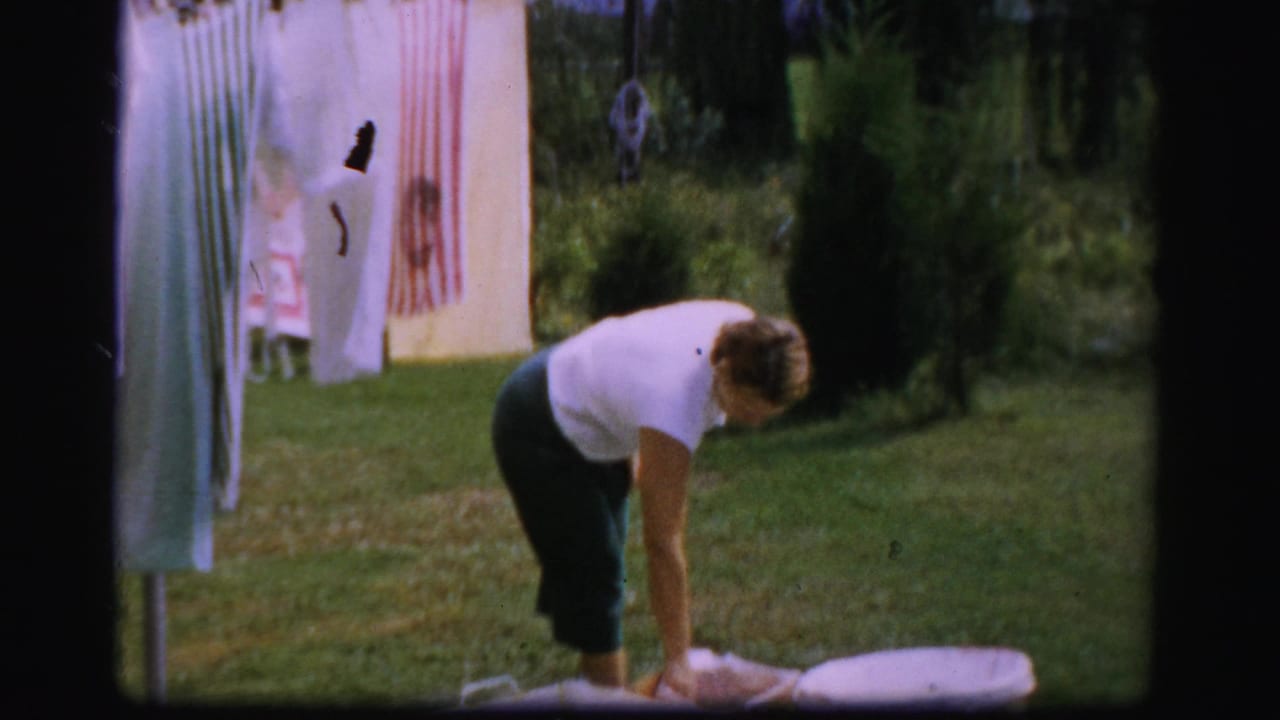 In Albany 1950s home-movie footage, a woman bends to handle laundry beside a rotary clothesline in a green backyard.