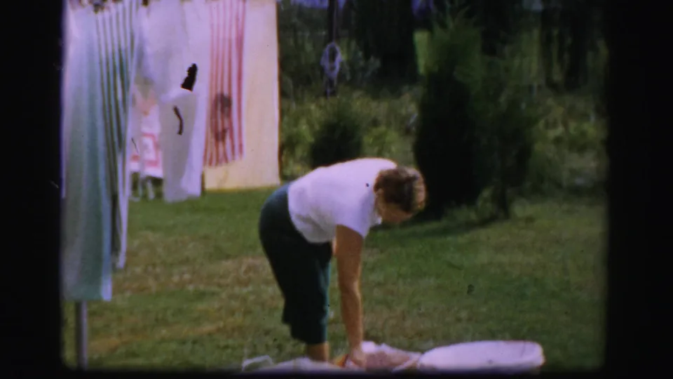 In Albany 1950s home-movie footage, a woman bends to handle laundry beside a rotary clothesline in a green backyard.