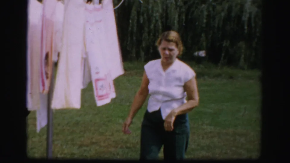 A woman walks past hanging white laundry in a grassy Albany backyard, captured with soft color 8mm home-movie texture.