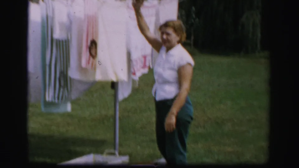 The woman reaches toward the hanging laundry on a rotary clothesline, with grass and trees visible in the Albany backyard.