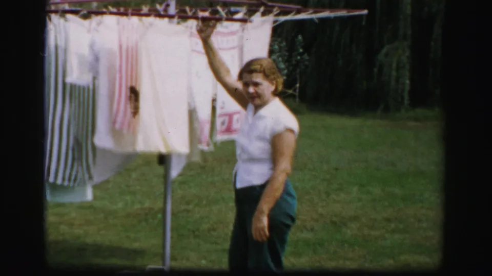 A woman in a white blouse reaches up to adjust laundry on a clothesline in an Albany yard, with grass and dark foliage behind her.