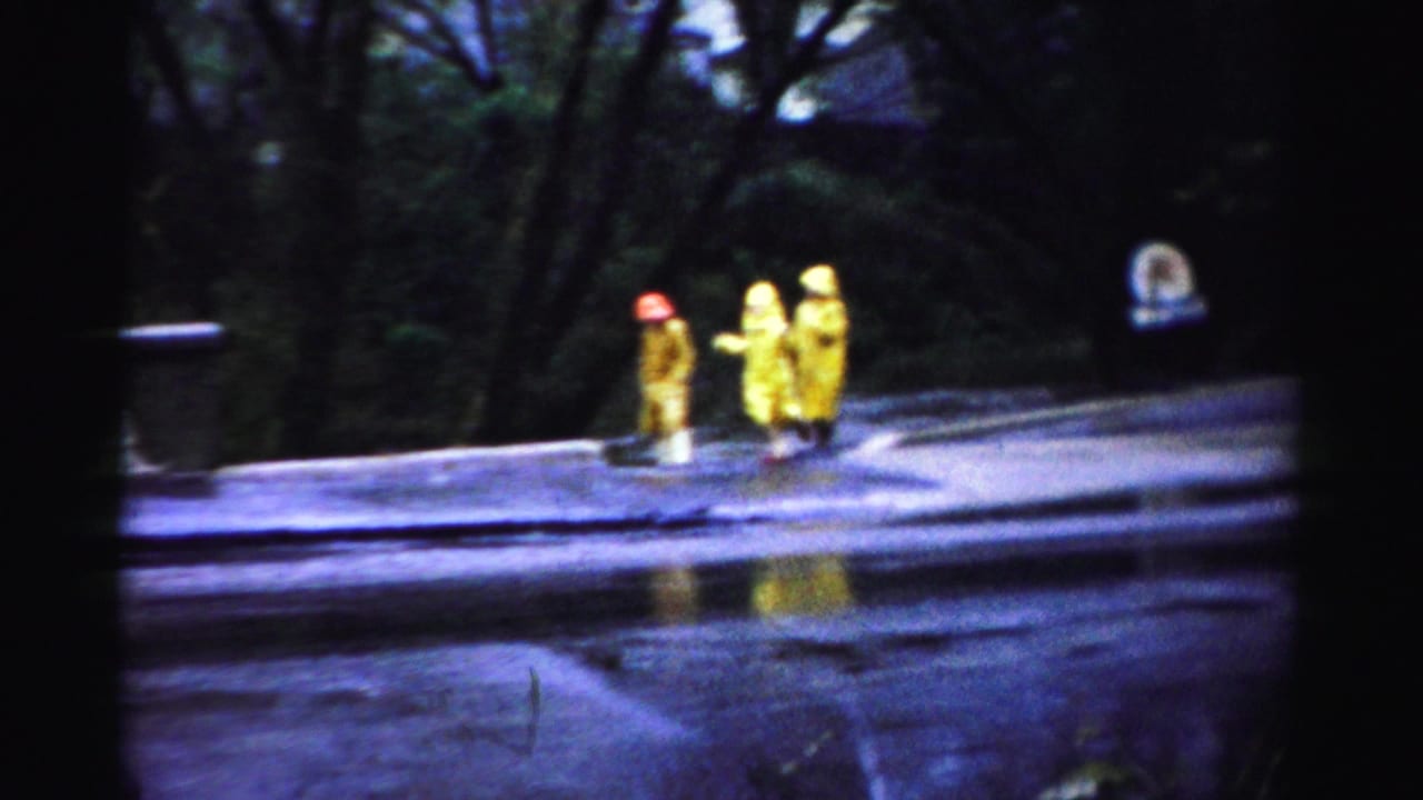 Albany-area 1950s 8mm frame showing two distant people in yellow rain gear on a wet roadside with dark trees behind them.