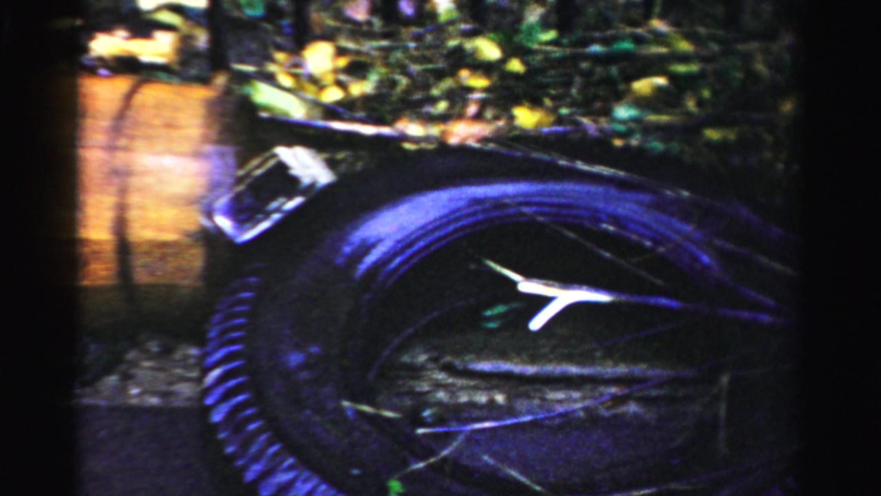 Albany, New York home-movie frame showing a dark car partly in floodwater near a wooden barrel and roadside debris.