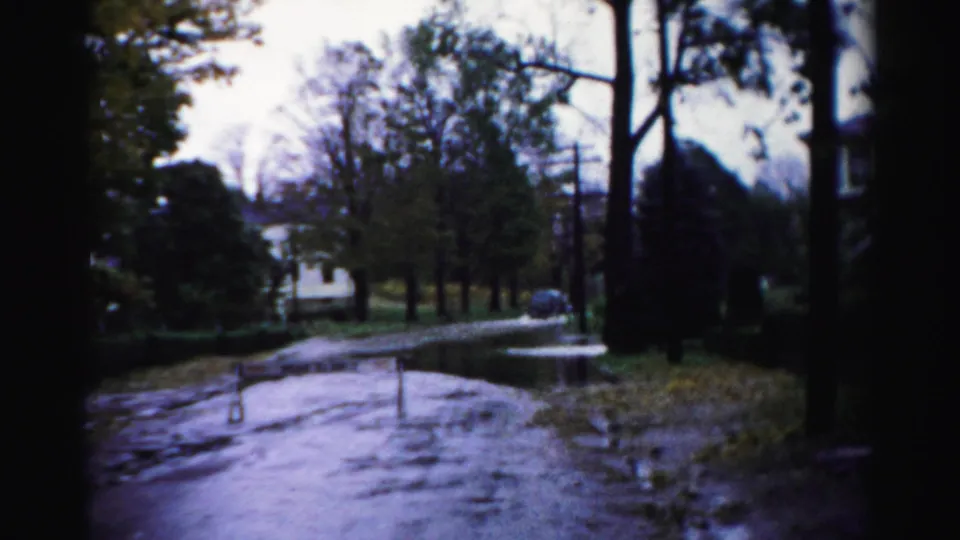 1950s Albany home-movie frame showing a flooded residential road bordered by trees, with a dark sedan near the far curb.