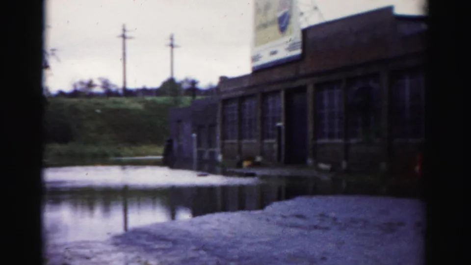 1950s Albany home-movie frame of a partially flooded commercial frontage, large rooftop or roadside sign, and reflective standing water.