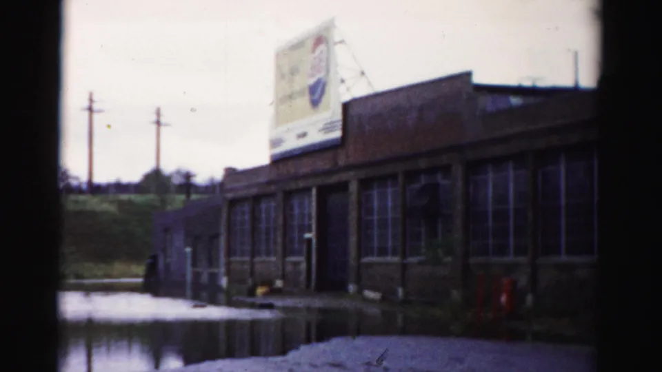 1950s Albany frame showing a low commercial building with many windows, a billboard-like sign, and standing floodwater along the frontage.