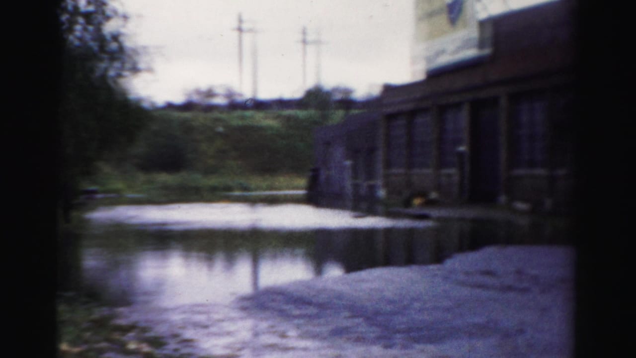 Floodwater covers a paved area near a brick structure, with a grassy bank and utility poles visible in the background.