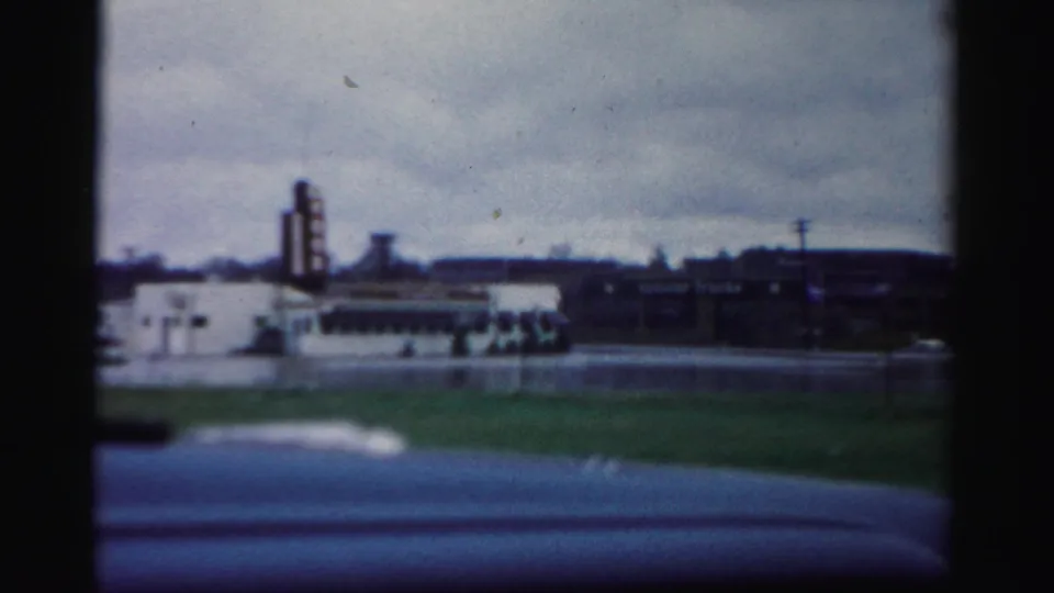 The frame shows flooded pavement and distant urban buildings, with utility poles and grass visible under a cloudy sky.