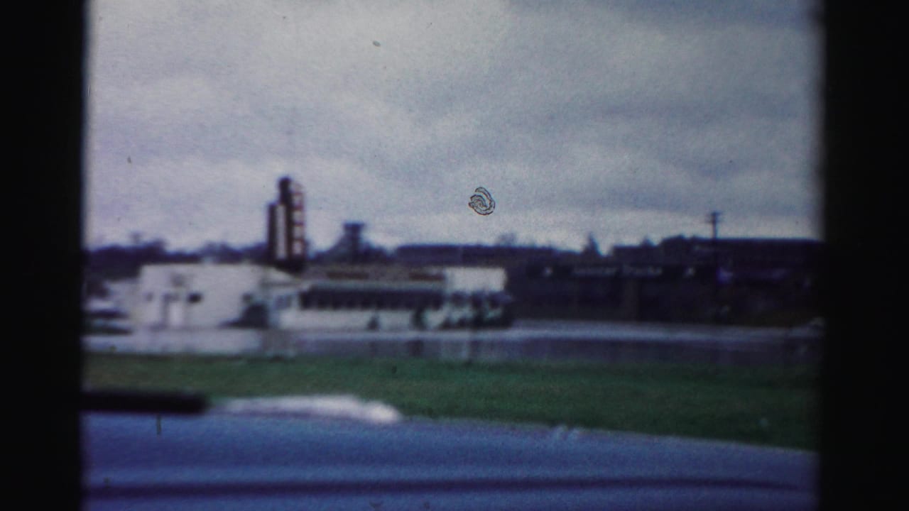 1950s Albany street footage showing wet pavement, grass, a distant industrial structure, and a gray overcast sky.