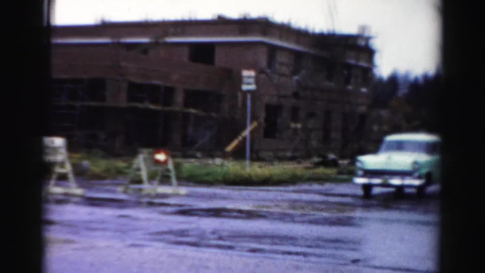 A 1950s Albany street scene shows a car near barricades and an unfinished brick building after rain.
