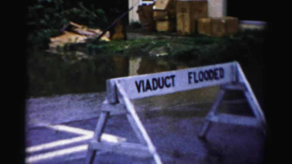 A roadside barricade reading VIADUCT FLOODED stands near pooled water, grass, and stacked materials in Albany.