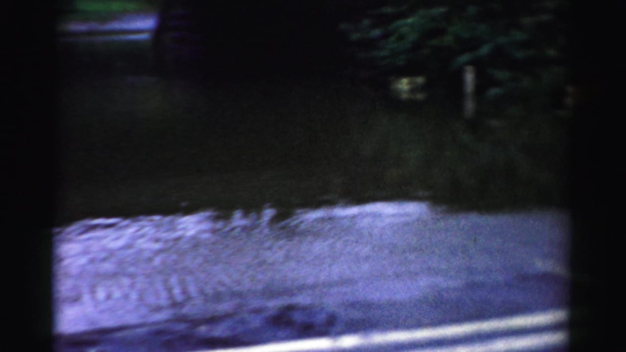 Albany 8mm frame with standing floodwater across a roadway, dark overhead structure, wet pavement, and dim reflected light.