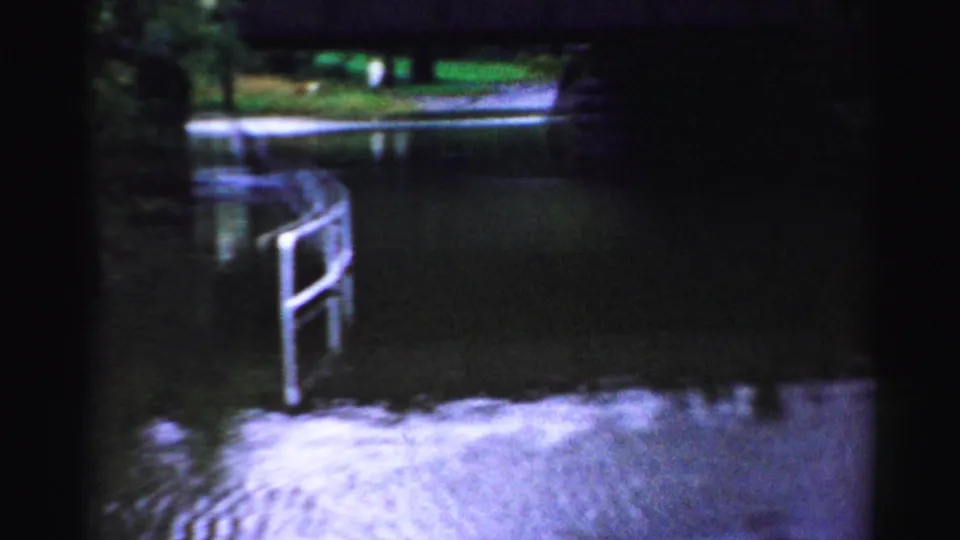 Albany 8mm frame showing a metal railing, standing water, wet pavement, trees, and reflections near the flooded viaduct approach.