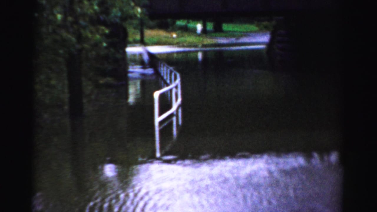 1956 Albany home-movie frame showing high water under a low bridge with a white railing partly submerged near the bank.