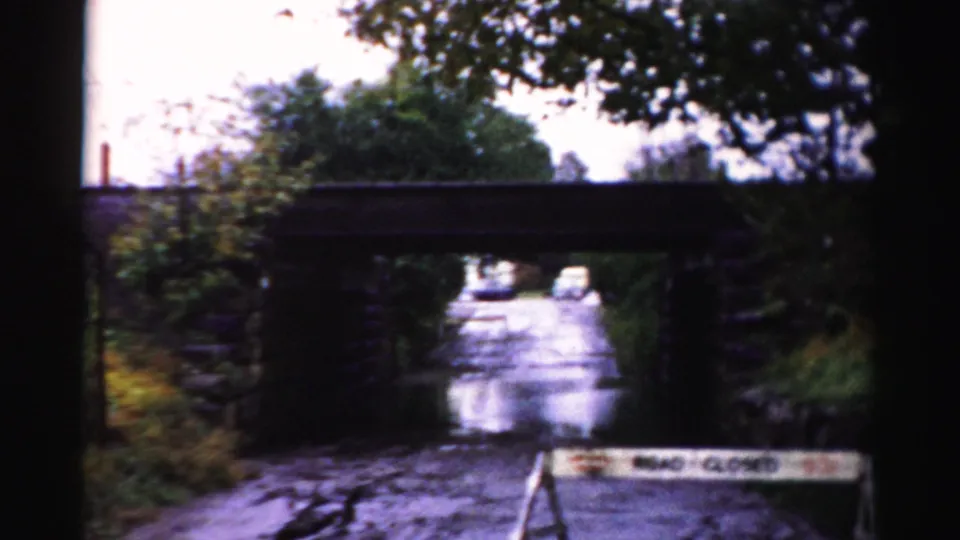 Wide 8mm view of a flooded Albany-area road under a stone bridge, with a partial ROAD CLOSED barricade and roadside trees.