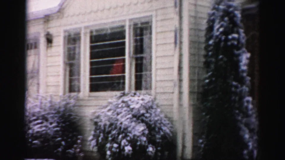 Brief winter home-movie frame showing a snow-dusted suburban house exterior, window, shrubs, siding, and evergreen tree.