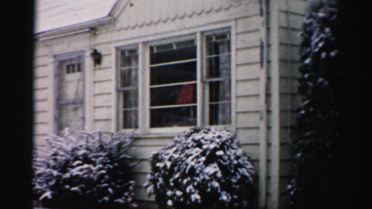 1956 Albany home-movie frame showing a snowy clapboard house exterior with shrubs, windows, and a small front entry.