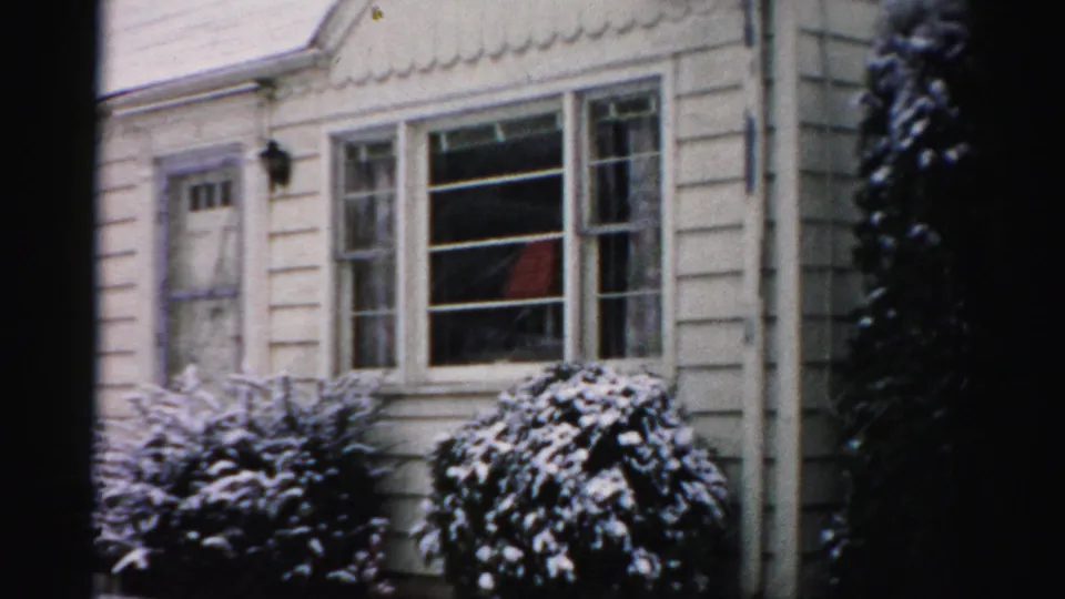 1956 Albany home-movie frame showing a snowy clapboard house exterior with shrubs, windows, and a small front entry.