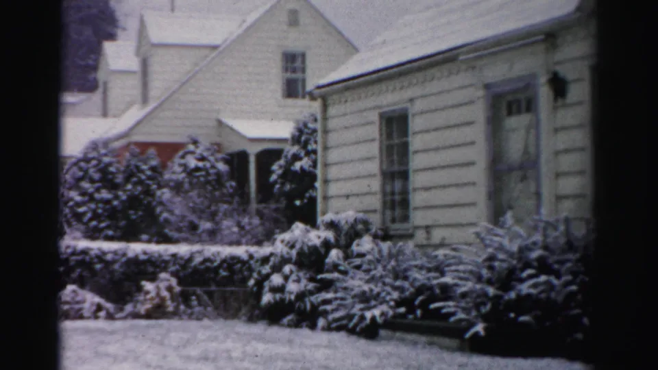 1950s Albany home-movie frame showing a white house, bare trees, and shrubs blanketed by fresh snow.
