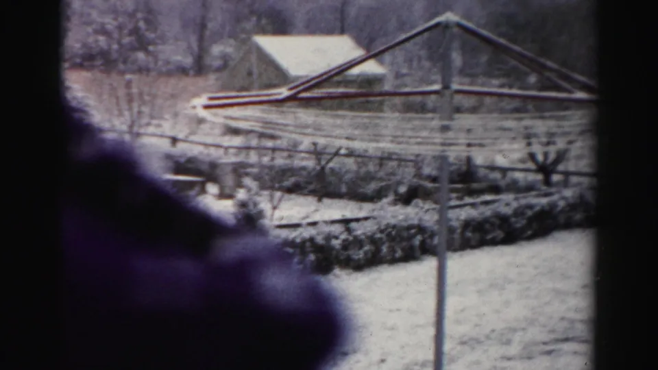 1956 Albany-area winter frame showing a clothesline pole in a snowy yard, with fencing, shrubs, bare trees, and a barn or shed roof behind it.