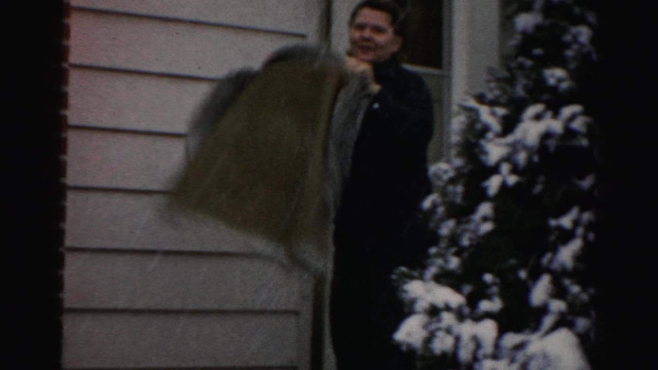 Adult woman on a snowy residential porch in the 1956 Albany-area home movie, standing beside light siding and evergreen shrubs.