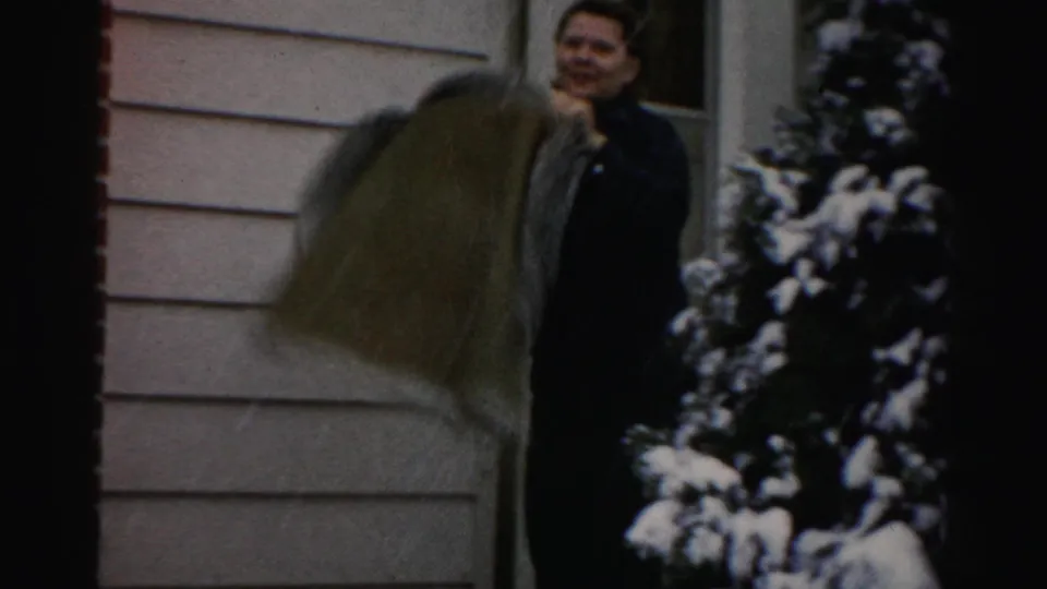 Adult woman on a snowy residential porch in the 1956 Albany-area home movie, standing beside light siding and evergreen shrubs.