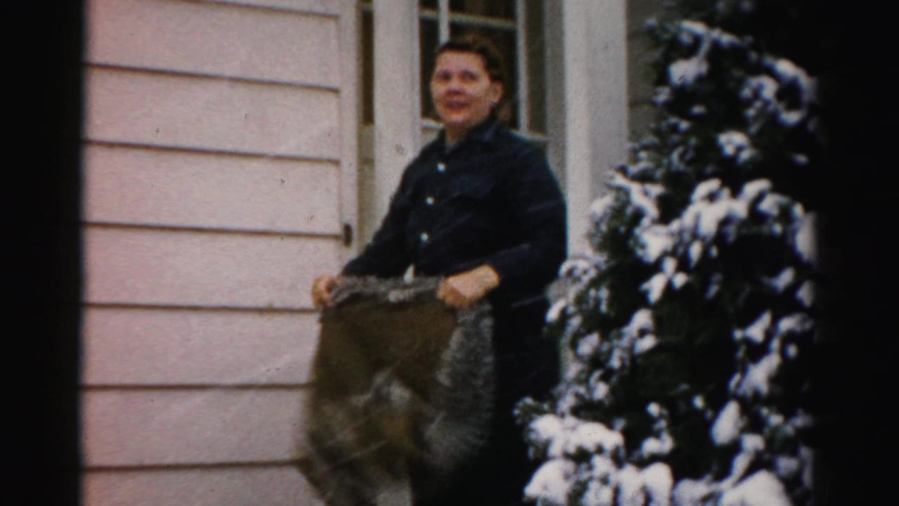 Woman in a dark coat holds a large blanket or rug outside a doorway beside snowy evergreens.