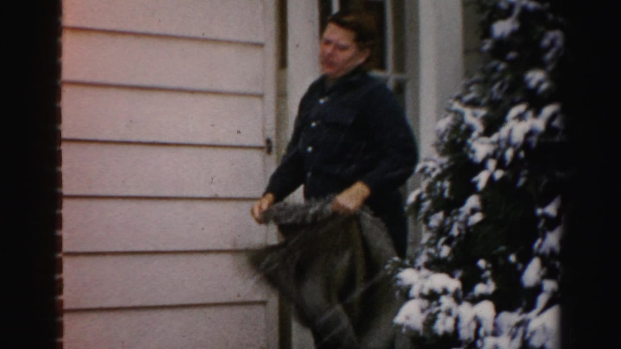 A woman in a dark coat holds a bulky blanket or rug outside a light-sided house in snowy Albany, New York footage.