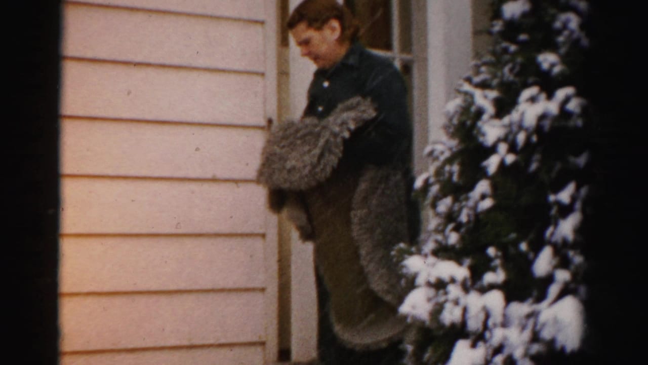 The woman gathers or shakes a heavy blanket or rug at the doorway, with pale siding and a snow-covered evergreen beside her.