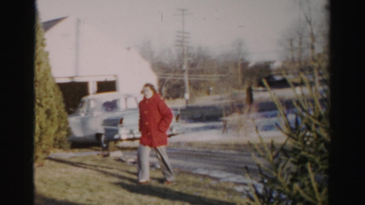 A woman in a red coat walks through an Albany yard near patches of snow, with a rounded 1950s car parked in the background.