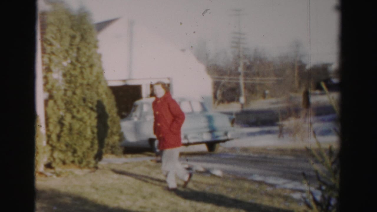 In Albany-area 1950s home-movie footage, a woman in a red coat crosses a winter lawn near a white house, driveway, garage, and pale car.