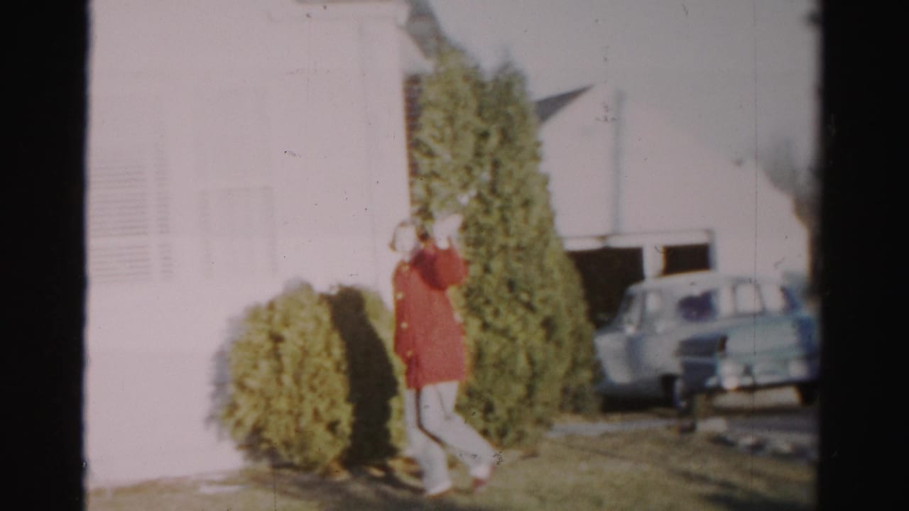Woman in a red coat waves while walking beside evergreen shrubs near a white house.
