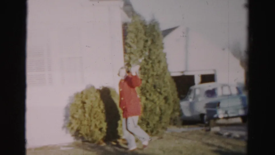 The woman waves toward the camera while passing evergreen shrubs and the white house exterior, with a pale car visible near the garage.