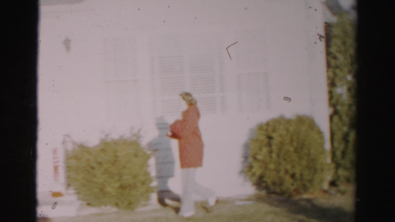 The woman nears the front door area of the Albany house, walking past low shrubs and white siding in soft, overexposed 8mm footage.